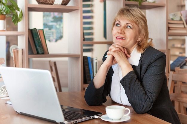 A woman in the office is working on a laptop. She smiles. A middle-aged woman, an adult in a cafe.