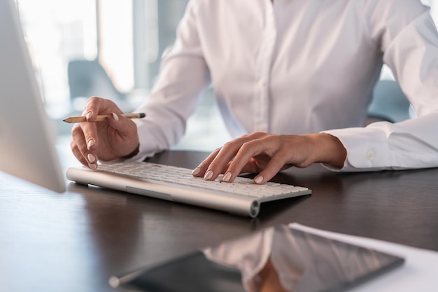 白いシャツを着た女性のオフィスマネージャーが白いキーボードのクローズアップにコンピューターの左手でテーブルに座っている顔なしオフィスワークの鉛筆の概念を保持している女性の手入れの行き届いた爪
