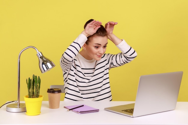 Woman office worker showing bunny ears with hands and smiling looking on laptop screen on video call