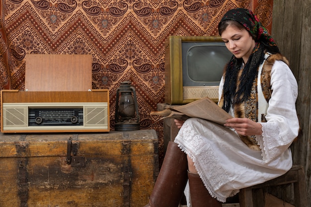 woman in old national clothes sitting on the chair among vintage room and reading newspaper, retro tv, radio and gas lamp on the wooden chest