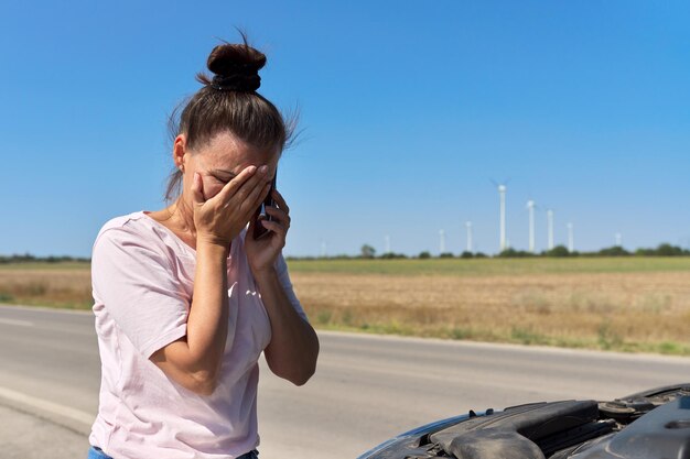 携帯電話で話している壊れた車の近くの道端の女性。悲しい女性は車の開いたボンネットを見て、専門的なサービスの支援を待っています