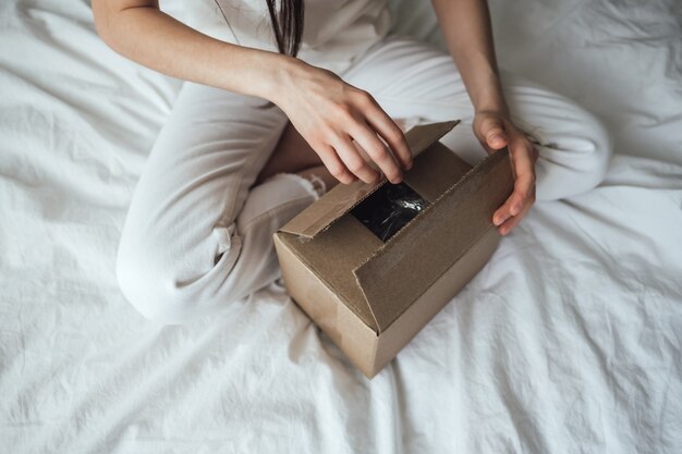 Woman opens a cardboard box, the parcel sitting on the bed at home. Delivery concept