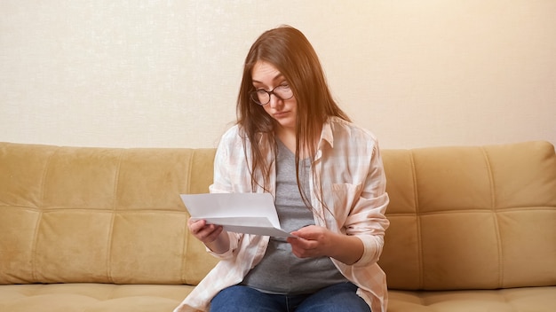 Женщина открывает конверт со счетом и нервничает на диване