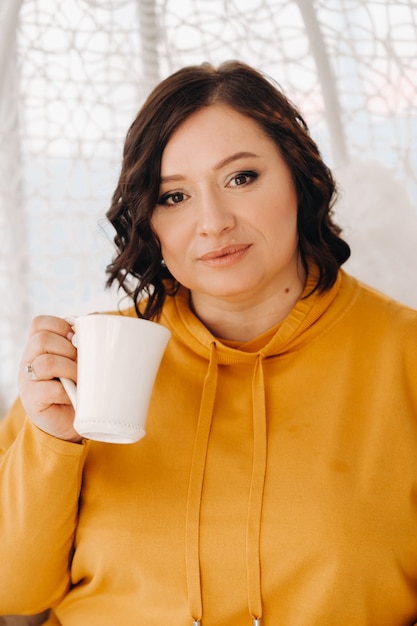 Una donna con una felpa arancione si siede su una sedia insolita e beve caffè a casa.