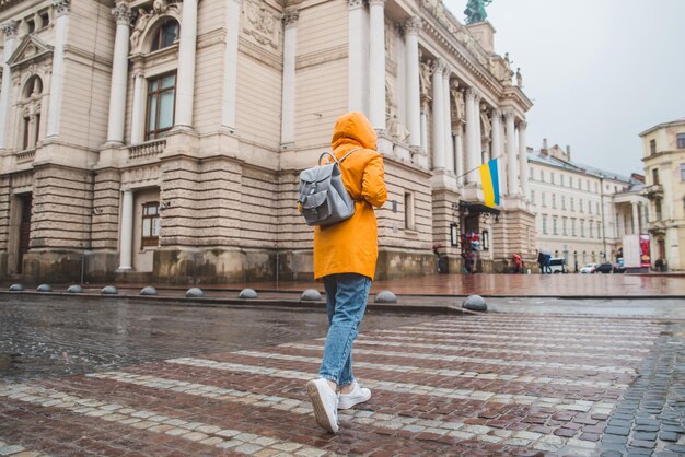 Женщина в оранжевом плаще пересекает дорогу старому европейскому зданию на заднем плане