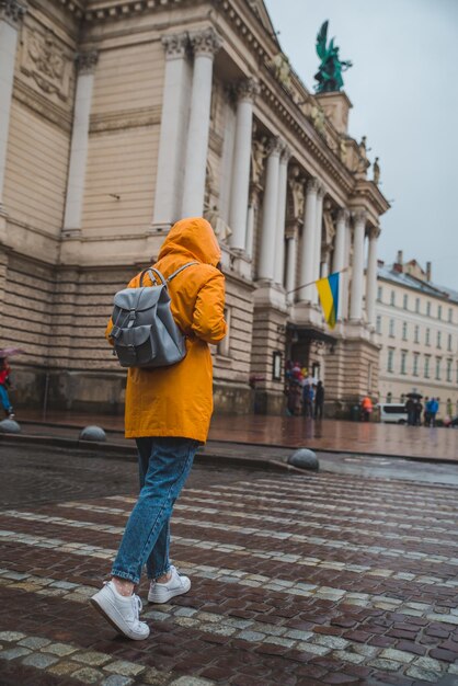 Женщина в оранжевом плаще пересекает дорогу старому европейскому зданию на заднем плане