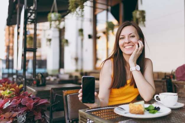 Donna in una caffetteria all'aperto di strada seduta al tavolo con una tazza di tè, torta, tenere in mano il telefono cellulare con schermo vuoto vuoto