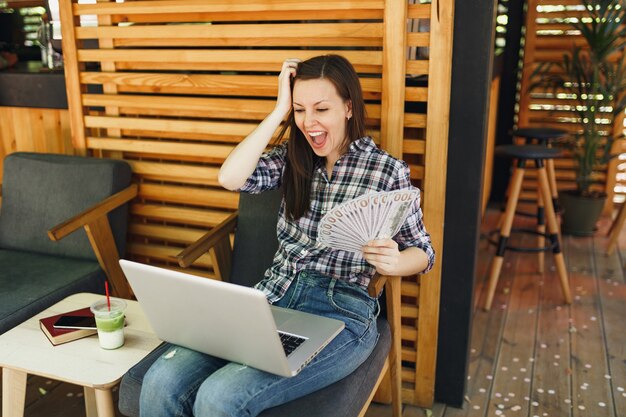 Woman in outdoors street coffee shop cafe sitting with modern laptop pc computer, hold in hand bunch of dollars banknotes, cash money