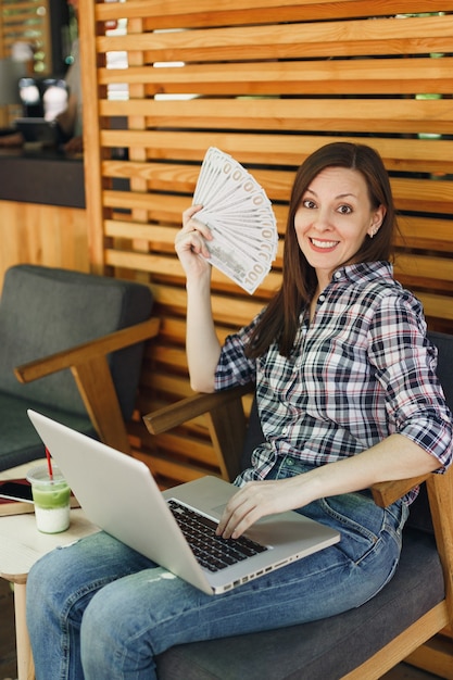 Woman in outdoors street coffee shop cafe sitting with modern laptop pc computer, hold in hand bunch of dollars banknotes, cash money