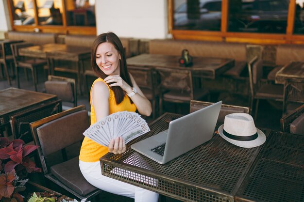 Woman in outdoors street coffee shop cafe sitting with modern laptop pc computer, holds in hand bunch of dollars banknotes