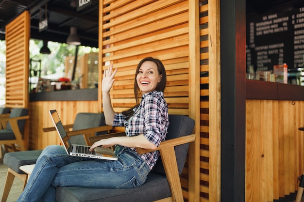 カジュアルな服に座って、現代のラップトップPCコンピューターで作業している屋外通り夏のコーヒーショップ木製カフェの女性