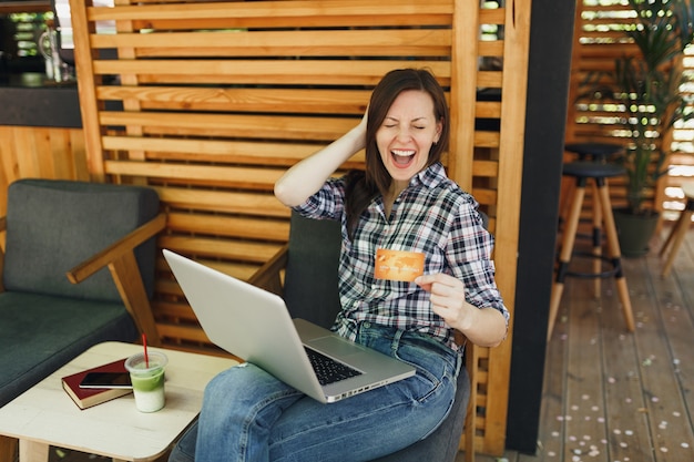 Donna in strada all'aperto caffè estivo caffè in legno seduto a lavorare su computer pc portatile, tenere la carta di credito bancaria rilassante durante il tempo libero