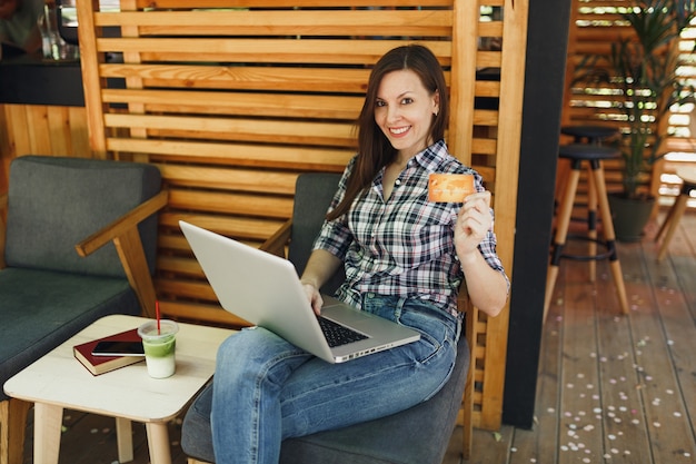 Woman in outdoors street summer coffee shop wooden cafe sitting working on laptop pc computer, hold bank credit card relaxing during free time