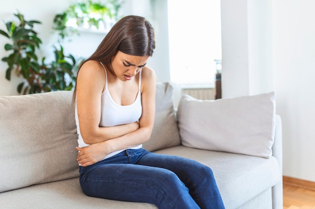 Woman in painful expression holding hands against belly suffering menstrual period pain, lying sad on home bed, having tummy cramp in female health concept