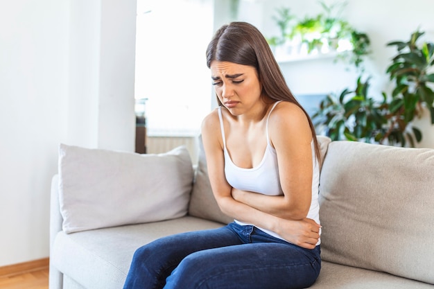 Woman in painful expression holding hands against belly suffering menstrual period pain, lying sad on home bed, having tummy cramp in female health concept