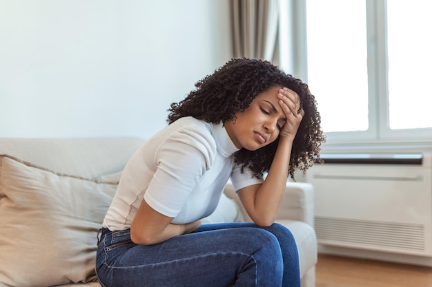 Woman in painful expression holding hands against belly suffering menstrual period pain, lying sad on home bed, having tummy cramp in female health concept