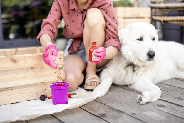 Женщина красит деревянный ящик, сидя с собакой на террасе