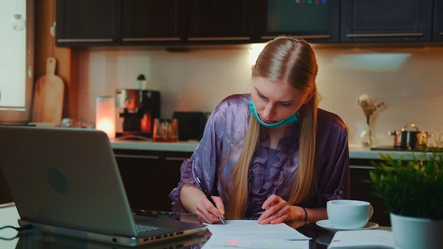 パジャマを着て、医療用マスクで書類に署名している女性は、キッチンのコンピューターで働いています...