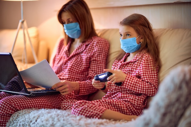 Woman in pajamas with notebook and papers working from home wearing protective mask while her kid, daughter playing computer console games