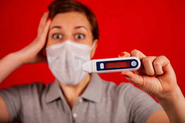 コロナウイルスCOVID-19感染によるパニック状態の女性。コロナウイルスと赤い背景の上のマスクを着た女性はCOVID-19の碑文と温度計で彼女の温度を読み取ります