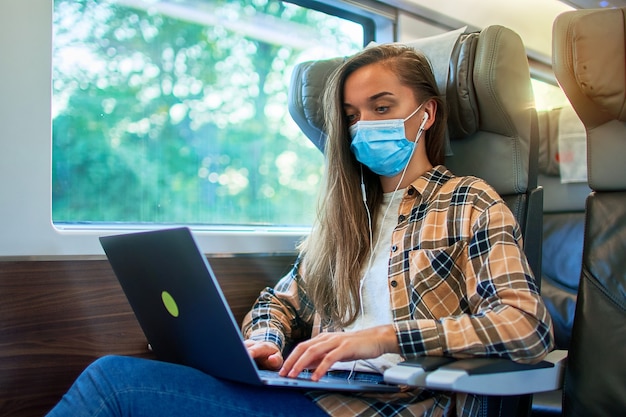 Woman passenger in face mask and headphones using a laptop for online working, watching video and reading news while safety travel by train. Virus protection and wearing mask in public transport