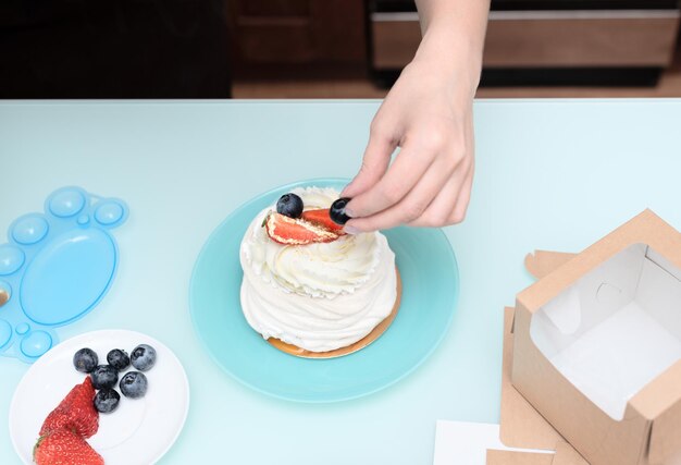 Woman pastry chef at home making finishing touches for homemade meringue cake Pavlova with summer fresh berries with selective focus Homemade pastry delicious sweets concept