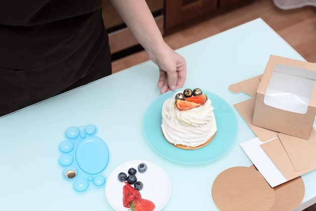 Woman pastry chef at home packs homemade meringue cake with summer fresh berries in eco craft box for delivery Cooking at home concept