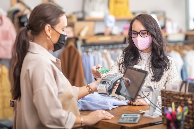 Woman pays for clothes contactless in the fashion store to the shop assistant who is holding a payment terminal, both wearing face masks.