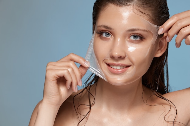 Woman peels-off transparent facial mask and smiling to the camera on blue