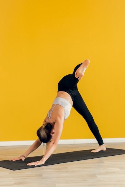 woman performs a difficult yoga asana, standing on a mat against a background of a yellow wall