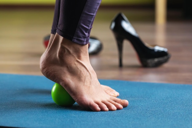 A woman performs a foot muscle massage with a massage ball\
while standing on a mat in the studio prevention of tired legs\
swollen veins after wearing shoes with heels