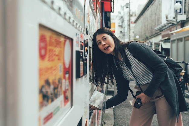 woman photographer buying drink coffee snack from automatic vending machine on japanese city urban street on sunny day. girl backpacker with camera smiling taking out her food from hole looking aside