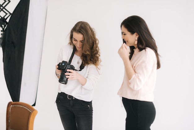 Woman photographer and female model together looking at made photo and talking,smiling, white background. Concept of creative work in photo studio, backstage job.