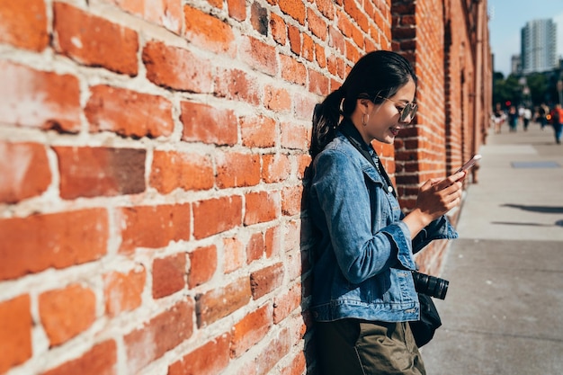 woman photographer traveling in san francisco city in summer holidays. young girl lens man using cellphone texting message online sending relying on the red brick wall under sunlight on busy street.