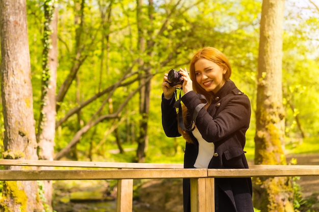 가을 공원에서 즐기는 검은색 재킷을 입은 여성 사진작가, 나무 다리에서 일몰 사진을 찍는다