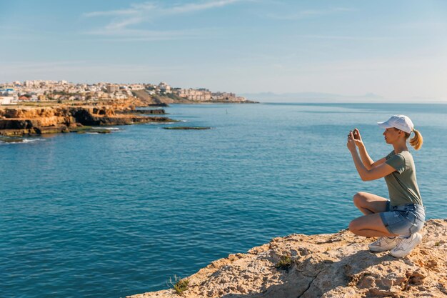 A woman photographs the city on a mobile phone by the sea