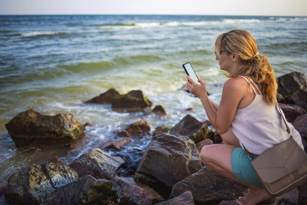 女性は石の上に座って電話で海の風景を撮影します