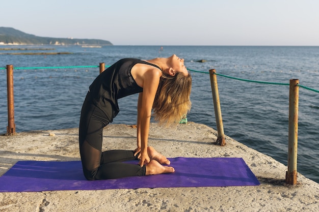 A woman on a pier on a sunny summer evening performs the ushtrasana exercise camel pose