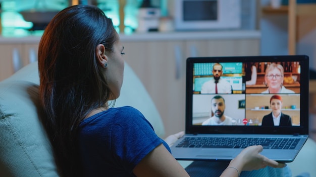 Woman in pijamas lying on sofa using laptop talking about sale report in video conference with team. Remote worker having online meeting consulting with colleagues using videocall and webcam chat