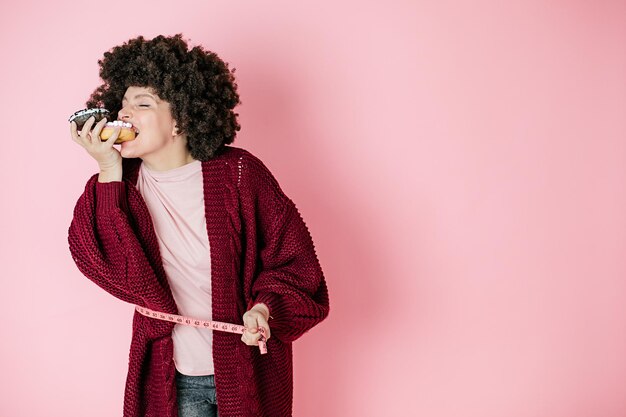 ピンクの背景の女性は、食欲をそそるドーナツ、髪型、アフリカンスタイルをかみます。彼女の手でそれを保持します。測定テープ。ユーモラスな写真。ダイエット。適切な栄養。