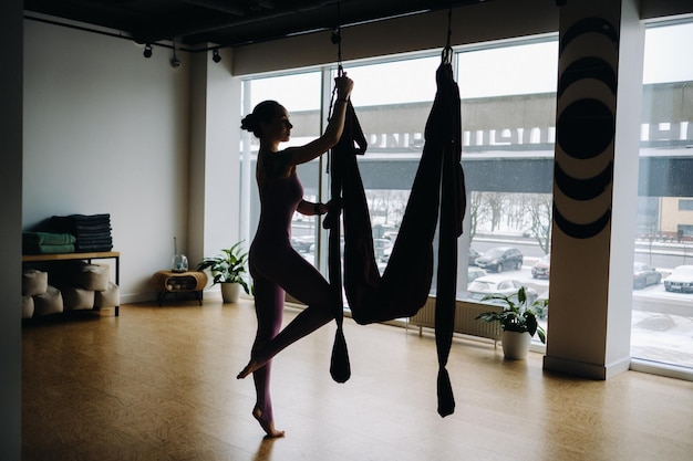 A woman in pink clothes does yoga on a suspended burgundy hammock in a bright gym