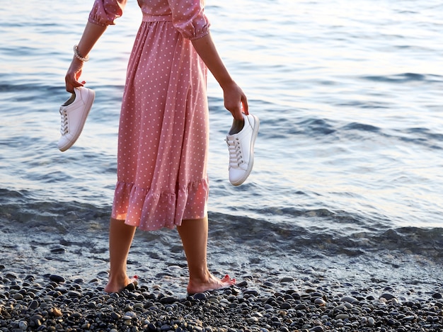 Woman in pink dress stands barefoot on edge of water on shore and holds her sneakers in his hands