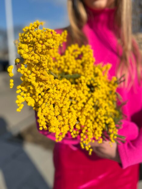 Женщина в розовой куртке держит букет желтых цветов.