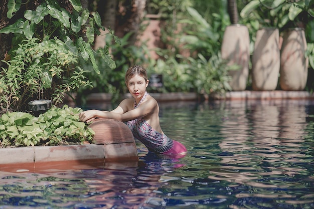 Женщина в розовом слитном купальнике расслабляется в тропическом бассейне, летние каникулы