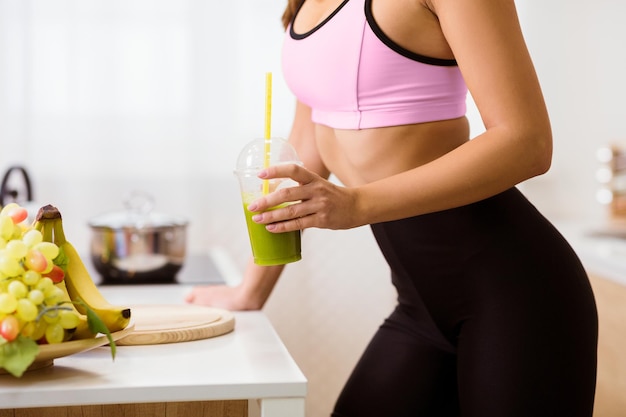 A woman in a pink sports bra and black leggings stands in a kitchen holding a green smoothie she has