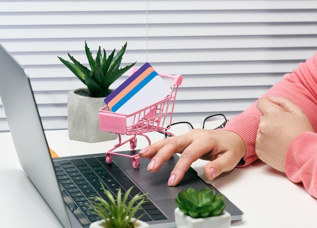 Woman in a pink sweater makes purchases online with a laptop, a credit card in hand. Woman sitting at a white table