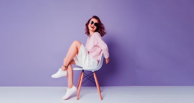 A woman in a pink sweater and white skirt sits on a chair in front of a purple wall.