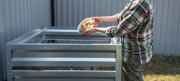 A woman in a plaid shirt pours food waste from a bowl into a compost heap of potato and carrot peelings Compost box made of metal ecofriendly fertilizer for the garden