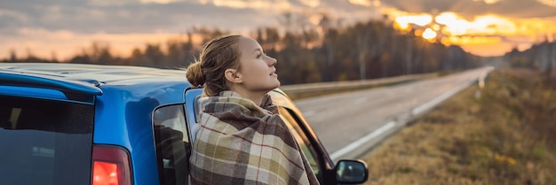 Женщина в пледе стоит у машины на обочине дороги на фоне рассветной поездки