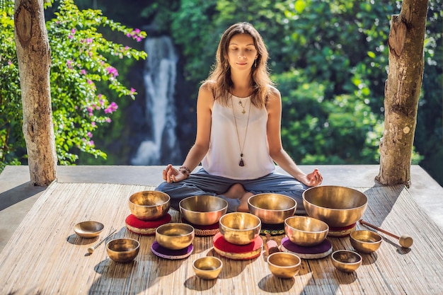 woman playing on tibetan singing bowl while sitting on yoga mat against a waterfall vintage tonned beautiful girl with mala beads meditating